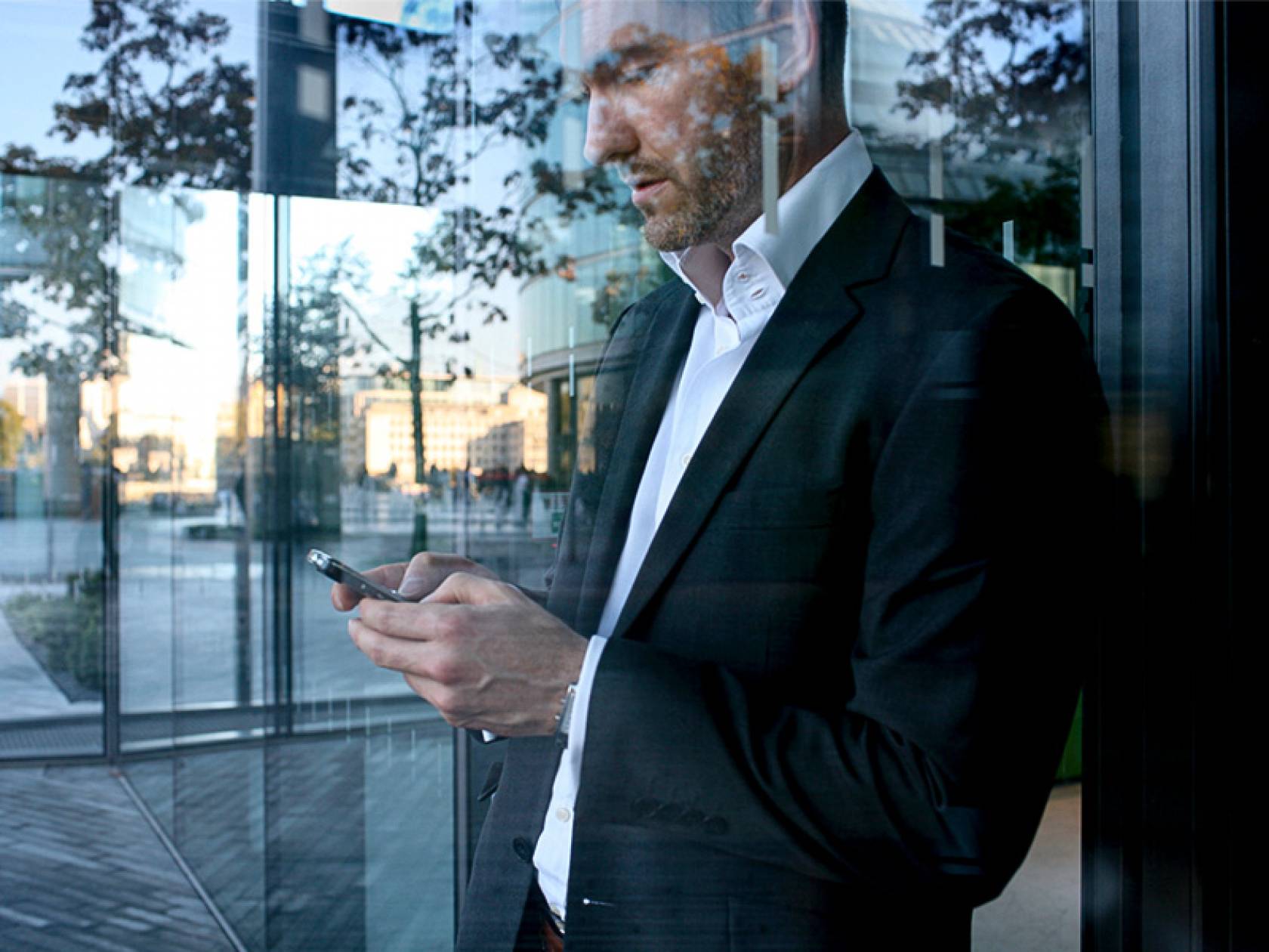 Schmuckbild Self Services und Apps Mann steht hinter einer Glasscheibe und schaut auf sein Smartphone